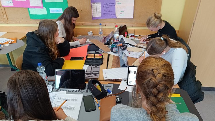 Sechs Schülerinnen sitzen an einem Tisch. Sie arbeiten mit Heften und Tablets. 