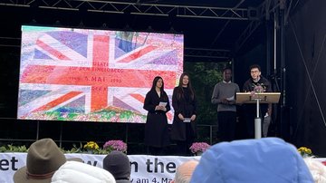 Zwei Schülerinnen und zwei Schüler auf der Bühne. Auf einer Leinwand die Britische Flagge.